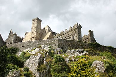 stone castle ruins atop a rocky hill, surrounded by greenery. the sky is cloudy, casting soft light on the ancient structures.