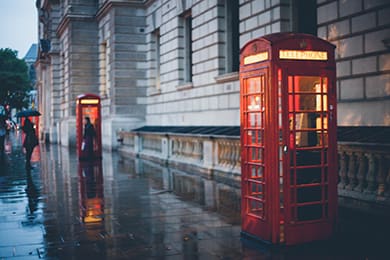 a rainy street scene featuring two iconic red british phone booths. the wet pavement reflects the city's lights. a person with an umbrella walks along the sidewalk beside the historic building.
