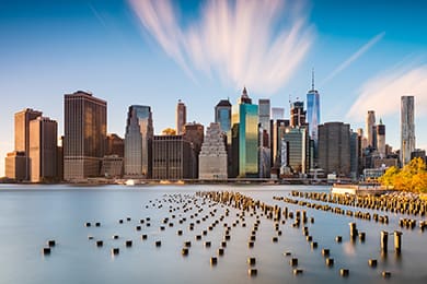 skyline of a city featuring tall buildings under a blue sky with streaks of clouds. in the foreground, a body of water with wooden pilings. trees are visible to the right. the scene is captured in warm, soft lighting.