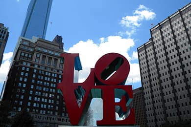 large red and blue "love" sculpture in an urban park setting with tall buildings and a blue sky with scattered clouds in the background.