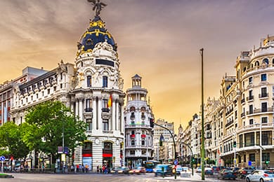 bustling city intersection with historical, ornate buildings. the prominent structure has a dark dome and decorative details. many people and vehicles are present, set against a vibrant evening sky. trees line the street, adding greenery.