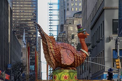 a large, colorful turkey float wearing a hat and bow tie is displayed in a city street surrounded by tall buildings. the float features bright, festive colors and stands out against the urban backdrop.