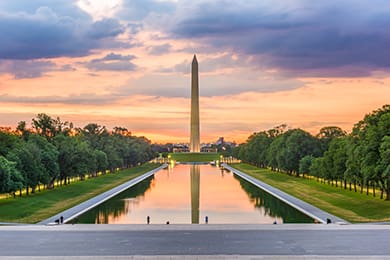 the washington monument stands tall at sunset, reflecting in the calm waters of the lincoln memorial reflecting pool. the sky is a mix of orange and purple hues, framed by lush green trees lining the pool.