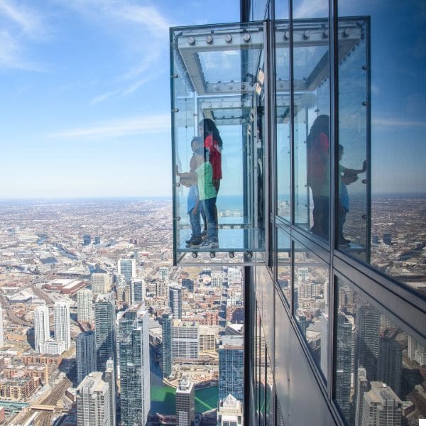 people stand inside a glass observation box extending from a skyscraper, overlooking a cityscape with numerous buildings under a clear blue sky. the box provides a dramatic view of the city below.