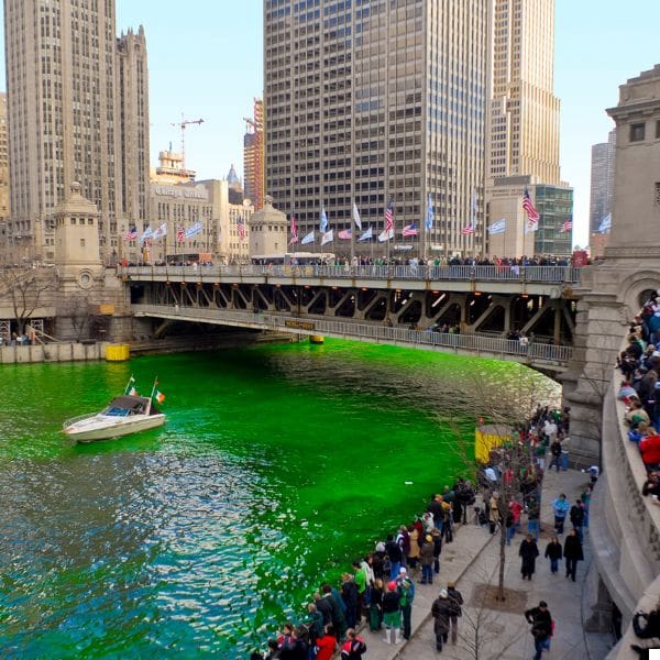 the chicago river dyed bright green for st. patrick's day, with a bridge and surrounding tall buildings. a boat sails on the river, and crowds of people line the riverbanks, watching the event. flags adorn the bridge above.