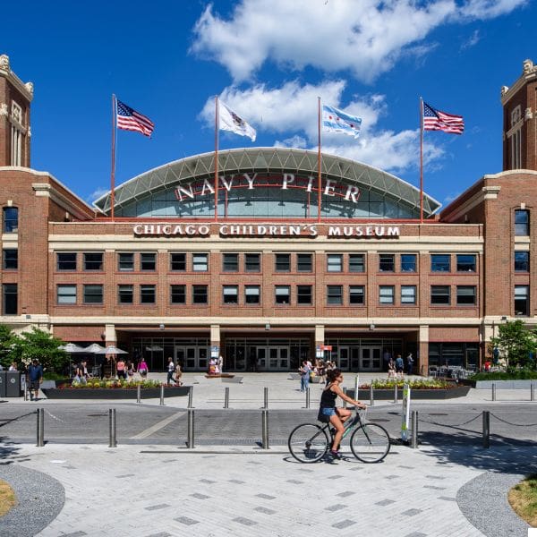 front view of navy pier in chicago under a blue sky with a few clouds. the brick building displays signs for the chicago children's museum. several flags are flying atop the building, and a person rides a bicycle in the foreground.