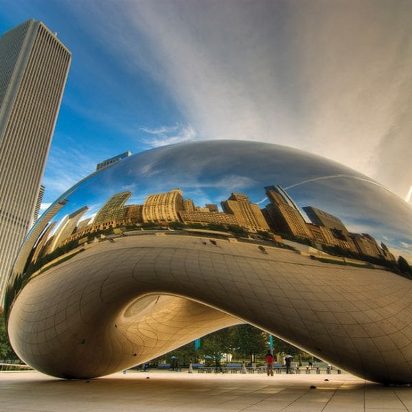 the image shows the reflective bean-shaped sculpture, cloud gate, in chicago’s millennium park. the skyline is mirrored on its surface under a blue sky with clouds.