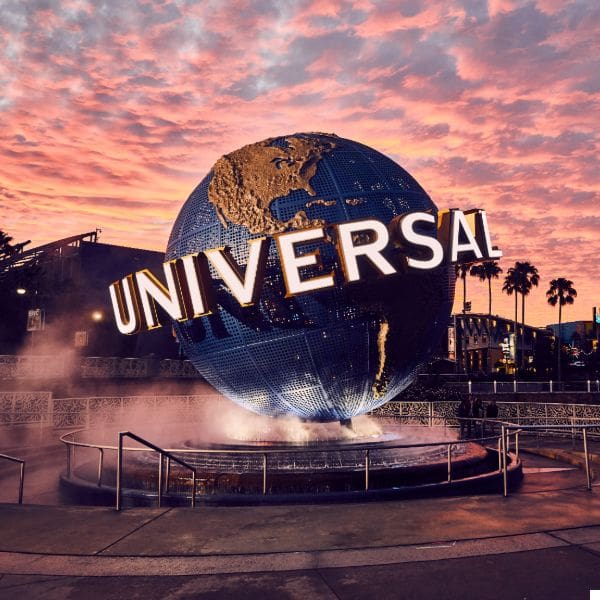 a large globe with "universal" written across it stands in the center of a plaza. the globe is surrounded by mist, and palm trees are visible in the background. the sky is vibrant with shades of pink and orange from a sunset.