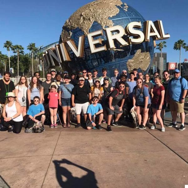 a large group of people pose together in front of the universal studios globe at the entrance. the globe features golden continents and the word "universal" in large letters. it's a sunny day with palm trees in the background.
