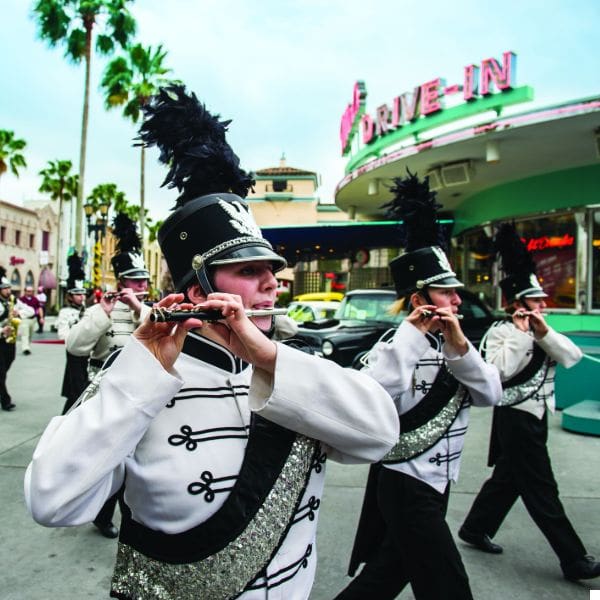 a group of marching band members in black and white uniforms play flutes as they walk past a retro drive-in diner. palm trees line the street, and classic cars are parked nearby.