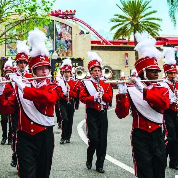 a marching band in red uniforms with white plumes plays instruments, including flutes and trumpets, while parading down a street. a roller coaster and palm trees are visible in the background.