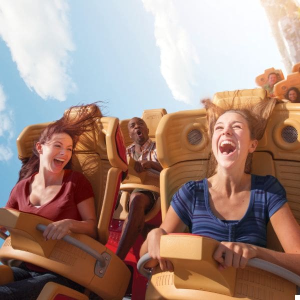 three people are riding a roller coaster, smiling and laughing with their hair flying in the air. the sky is clear and blue in the background, enhancing the sense of excitement and thrill.