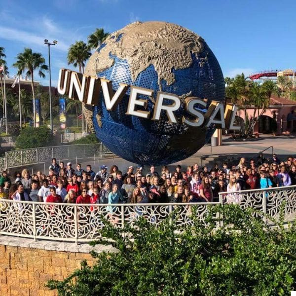 a large group of people pose in front of the iconic universal studios globe. the globe features a metallic map design with "universal" written across it. palm trees and park structures are visible in the background under a clear blue sky.