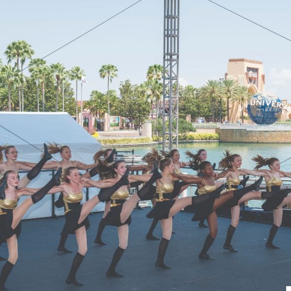 a dance group performs on an outdoor stage, each member wearing a gold top, black shorts, and matching knee-high socks. they all have one leg raised high in unison. palm trees and a theme park globe can be seen in the background.