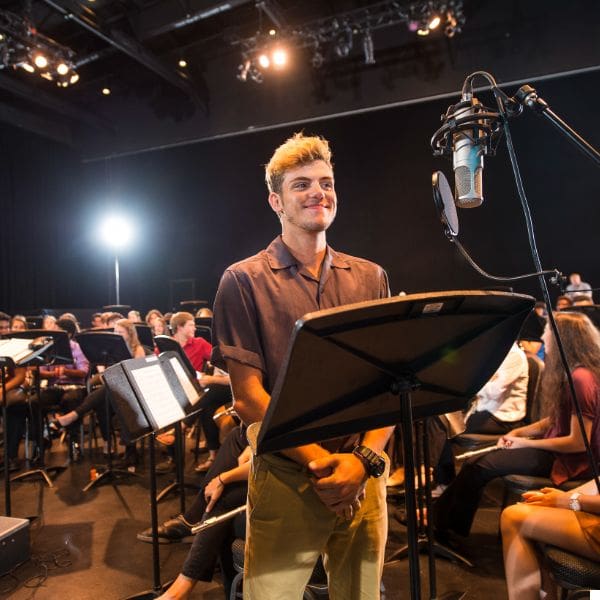a person stands at a microphone, smiling, in a dimly lit room. behind them, a group of people sit with music stands, suggesting a rehearsal or recording session. the atmosphere is focused and professional with studio lighting.