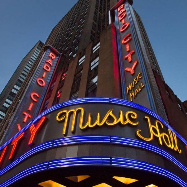 neon signs illuminate the facade of radio city music hall. the marquee features blue, red, and yellow lights, highlighting the words "radio city" and "music hall" against a backdrop of tall buildings in the evening.