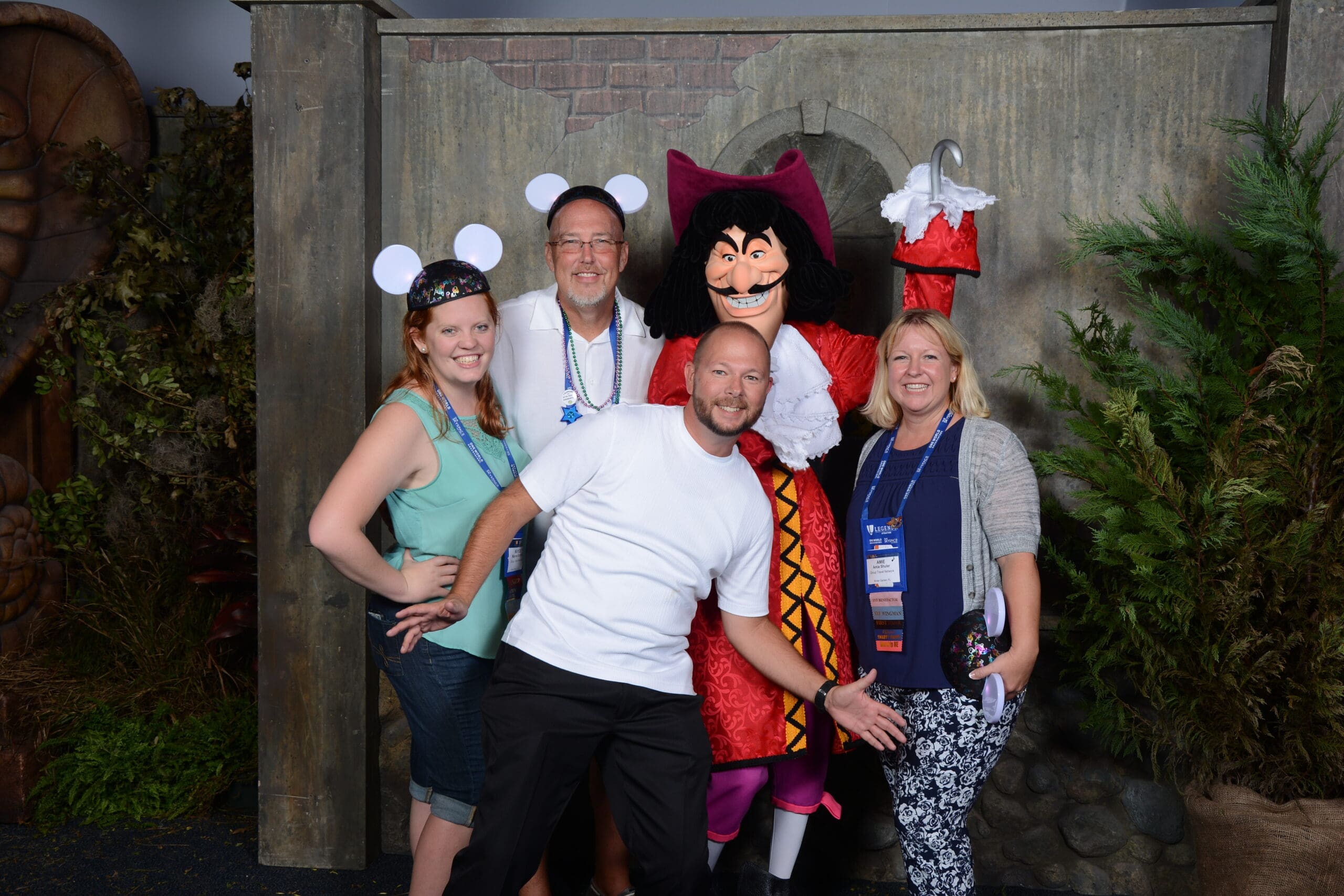 a group of four adults poses cheerfully with a captain hook character. one woman wears mickey mouse ears, and the man in front playfully spreads his arms. they all smile, standing in a themed setting with greenery and stone walls.
