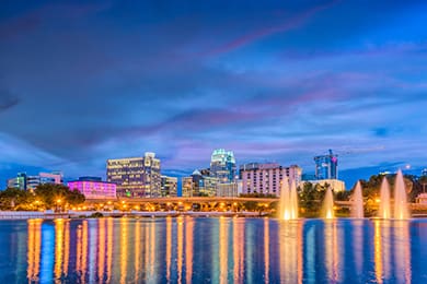 the orlando skyline at dusk showcases illuminated buildings reflecting off a tranquil lake. fountains inject dynamic movement, while a vibrant sky transitions from blue to pink, epitomizing this enchanting destination.