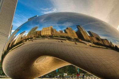 the image shows the cloud gate sculpture, also known as "the bean," in chicago's millennium park. the shiny, reflective surface mirrors the surrounding skyscrapers and blue sky, with a distorted view of the buildings and plaza.