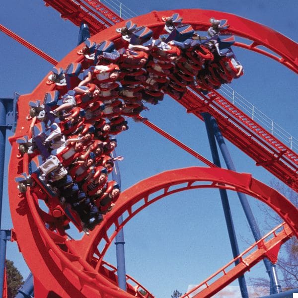 destination san antonio 6 people riding a roller coaster in a vertical loop. the track is red, and the sky is clear blue. riders appear excited, with some holding their hands up.
