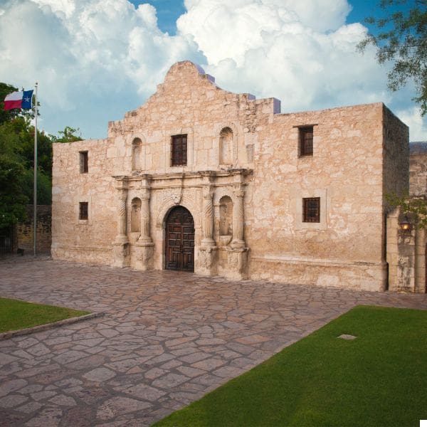 destination san antonio 5 a historic stone building with a weathered facade stands against a backdrop of fluffy clouds. a flag flies on a pole to the left. the foreground features a spacious stone courtyard and green lawns.