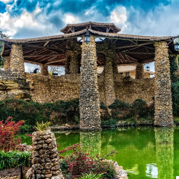 destination san antonio 3 a rustic stone pavilion with a thatched roof stands over a lush green pond. the pavilion is supported by multiple stone pillars and features hanging lanterns. surrounding foliage and a cloudy sky add to the serene atmosphere.