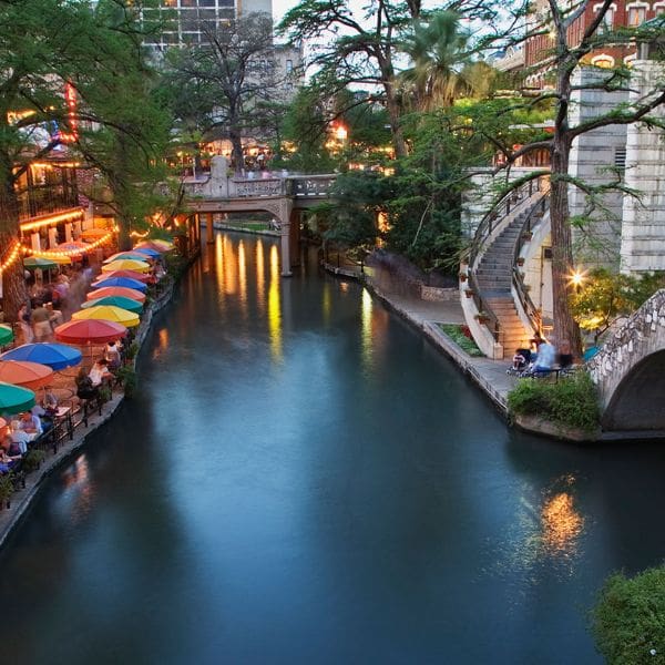 destination san antonio 4 scenic view of a vibrant riverwalk in san antonio, texas. colorful umbrellas line the riverside patios, with trees and historic buildings in the background. dimly lit with ambient lights, a peaceful atmosphere covers the winding waterway and bridges.