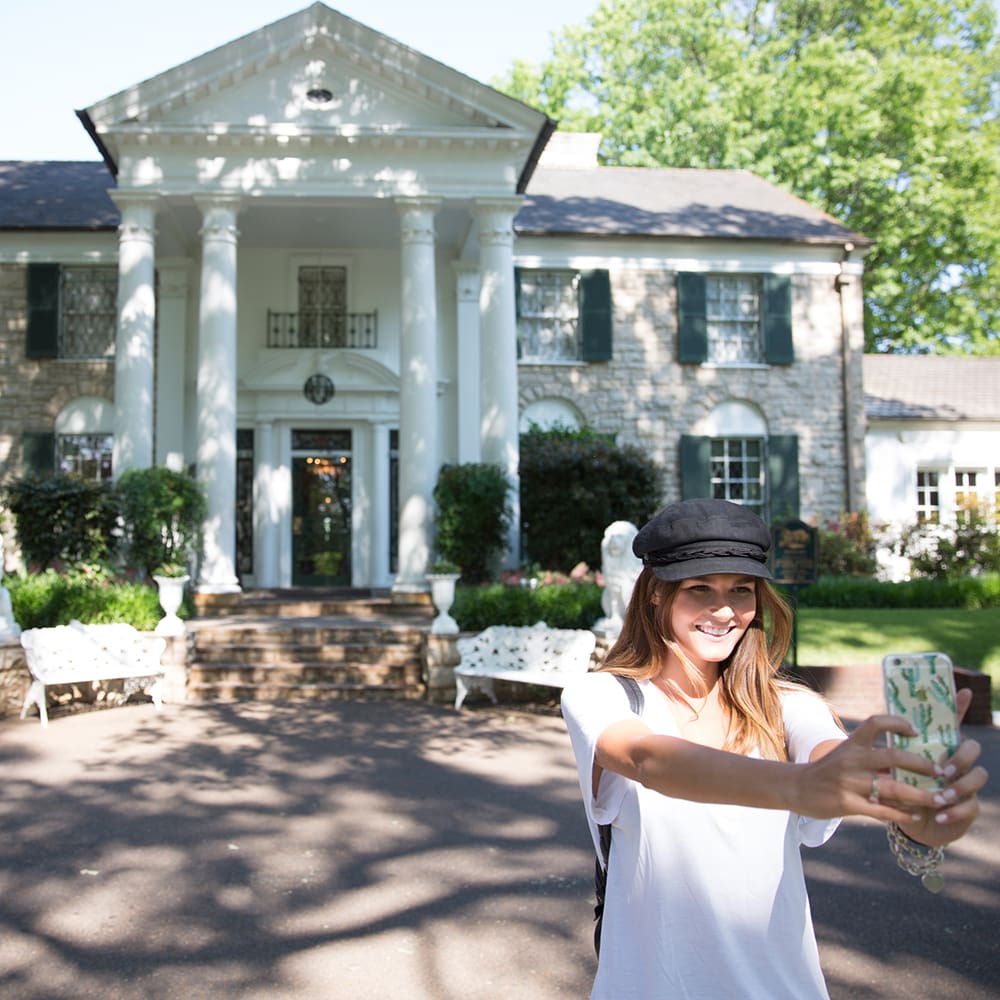 destination memphis 4 a person wearing a black cap and white shirt takes a selfie in front of a grand white-columned house with green shutters, surrounded by trees and a sunny sky.