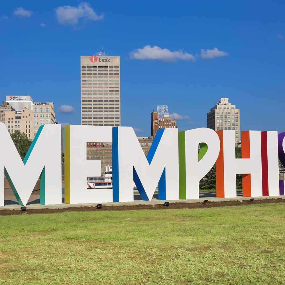 destination memphis 5 colorful large letters spelling "memphis" on a grassy area, with tall buildings and a clear blue sky in the background.