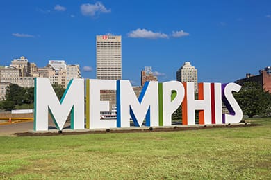 large white letters spelling "memphis" proudly stand as a landmark destination on a grassy area in front of a cityscape under a clear blue sky. the letters boast colorful edges, while tall buildings tower in the background, capturing the vibrant spirit of this iconic city.