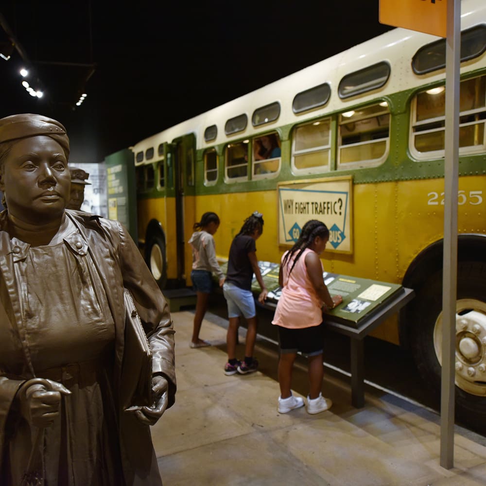 destination memphis 6 a bronze statue of a woman stands in the foreground. in the background, children interact with displays beside a vintage yellow and green bus. the bus has "why fight traffic? go by bus" written on its side.
