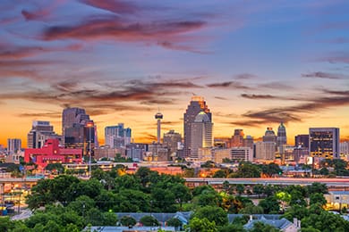 san antonio's vibrant city skyline at sunset features numerous tall buildings. the sky is painted with colorful hues of orange, pink, and purple. in the foreground, lush green trees create a striking contrast with this popular destination's urban landscape.