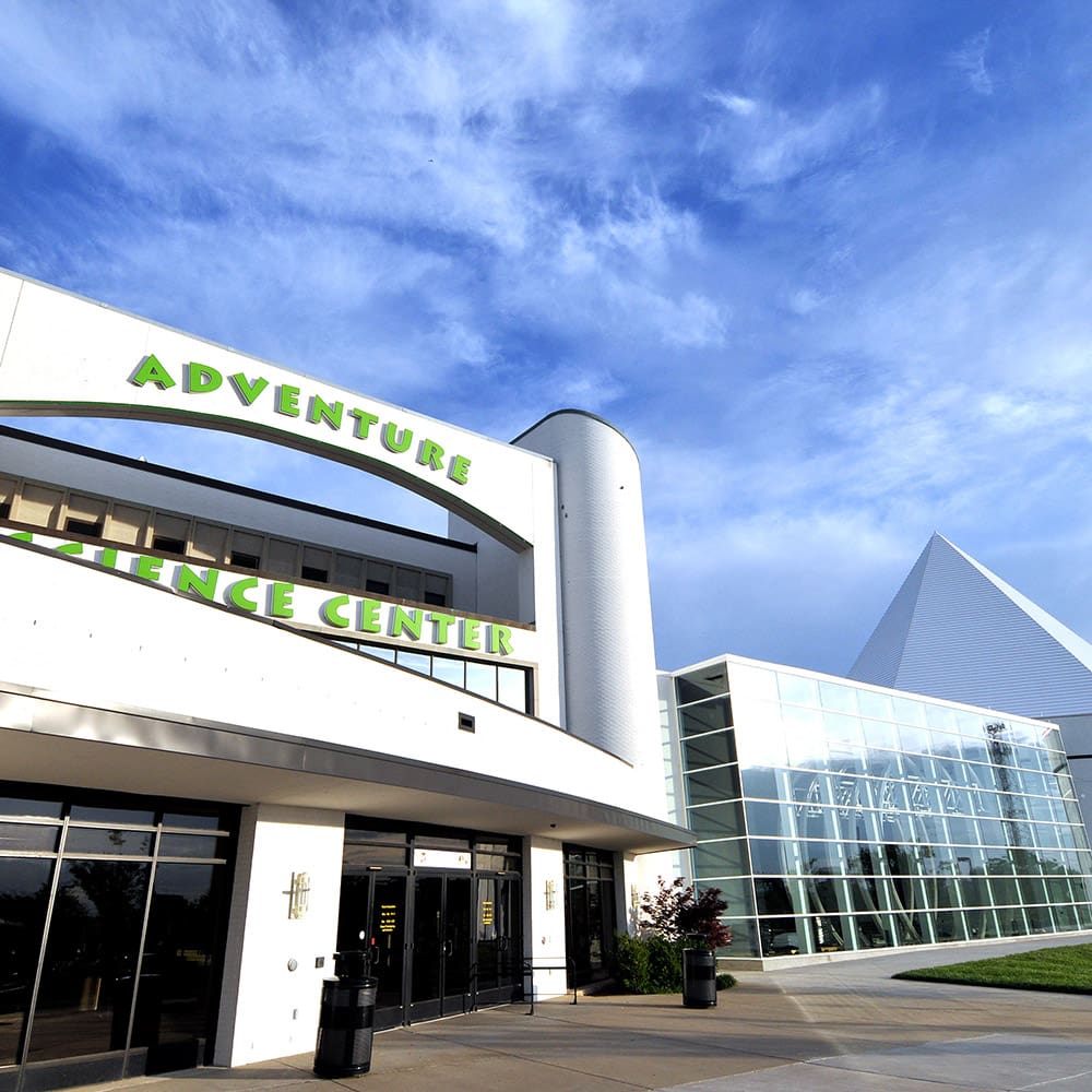 destination nashville 6 front view of the adventure science center building, featuring modern architecture with a cylinder and pyramid shape, large glass windows, and a blue sky backdrop. the building's name is displayed prominently in green letters.