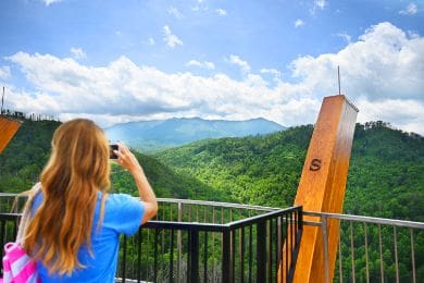 woman in blue shirt takes a photo of lush green mountains from a viewing platform with wooden beams. bright blue sky and clouds enhance the scenic background.