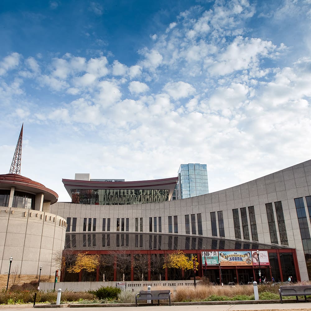 destination nashville 5 exterior view of a modern museum with curved architecture under a partly cloudy sky. the building features large windows and banners near the entrance, with some autumn trees nearby.