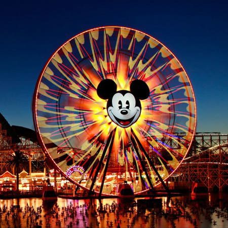 destination los angeles & southern california 4 a ferris wheel at dusk with bright, colorful lights, featuring a large, smiling mickey mouse face at its center. the night sky is clear, and the lights reflect on the water below, creating a festive atmosphere.