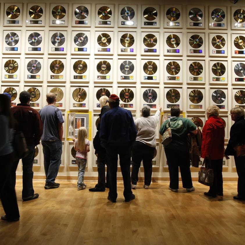 destination nashville 3 a group of people stand in front of a wall displaying numerous gold records in frames, lined up in rows. the floor is wooden, and the lighting is focused on the records, highlighting their gleaming surfaces.