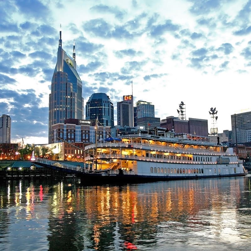 destination nashville 7 a riverboat is docked on a calm river reflecting city lights at dusk. the skyline features modern skyscrapers against a partly cloudy sky, with lights illuminating the buildings.