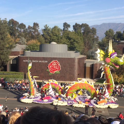 destination los angeles & southern california 5 a colorful dragon-shaped float adorned with flowers is parading down a street. crowds line the street, and the norton simon museum is visible in the background with trees and mountains on a clear day.