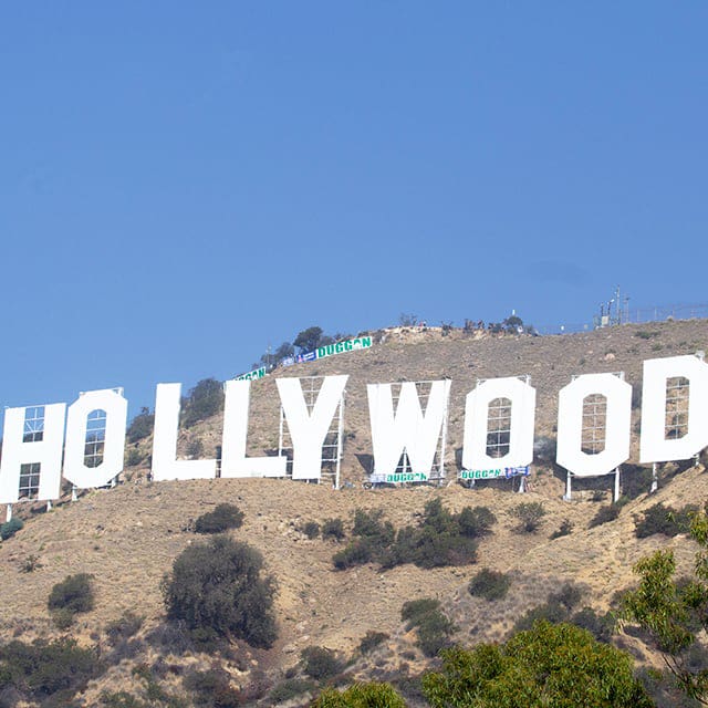 destination los angeles & southern california 3 the image shows the iconic hollywood sign in los angeles, set against a clear blue sky. the large white letters spell "hollywood" and are situated on a hilly, dry landscape with some greenery in the foreground.