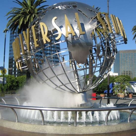 destination los angeles & southern california 6 a large metal globe with the words "universal studios" mounted on it, surrounded by mist and fountains. palm trees and tall buildings are visible in the background under a clear blue sky.