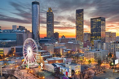 a cityscape of atlanta at sunset features illuminated skyscrapers and a glowing ferris wheel in the foreground. a colorful sky with clouds adds to the scene's vibrancy, highlighting the city's dynamic architecture and lively atmosphere.