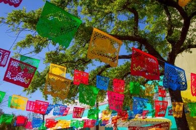 colorful papel picado banners hang beneath a tree with green leaves against a blue sky. the banners, in vibrant shades of red, blue, yellow, and green, sway gently, creating a festive atmosphere.