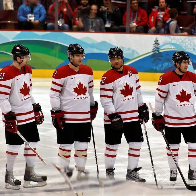 four hockey players in matching red and white team canada jerseys with maple leaf logos stand on the ice. they hold hockey sticks and wear helmets. the audience is in the background.