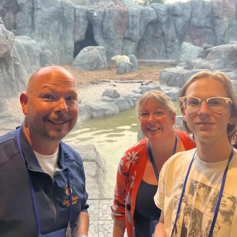 three people are smiling in the foreground with a rocky zoo exhibit and a polar bear in the background. they appear to be enjoying a day out. the setting is an outdoor, naturalistic animal enclosure.