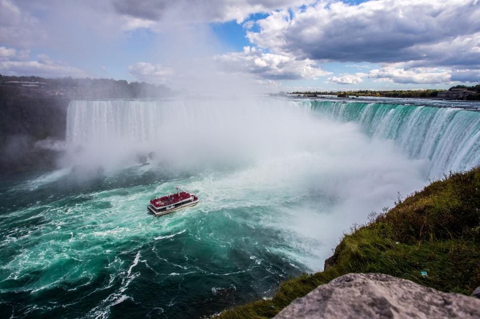 a small boat navigates near the base of a large waterfall, surrounded by mist and churning turquoise water. the waterfall spans a wide cliff, with a cloudy sky above. a rocky foreground and lush greenery are visible on the right.