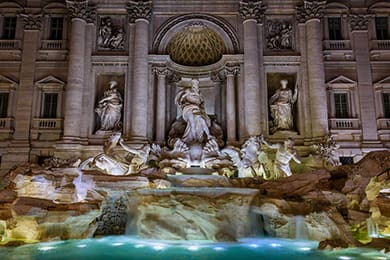 night view of the trevi fountain in rome, italy, illuminated with lights. the baroque structure features statues, including a central figure and horses with a cascading water pool in the foreground, set against an ornate facade.