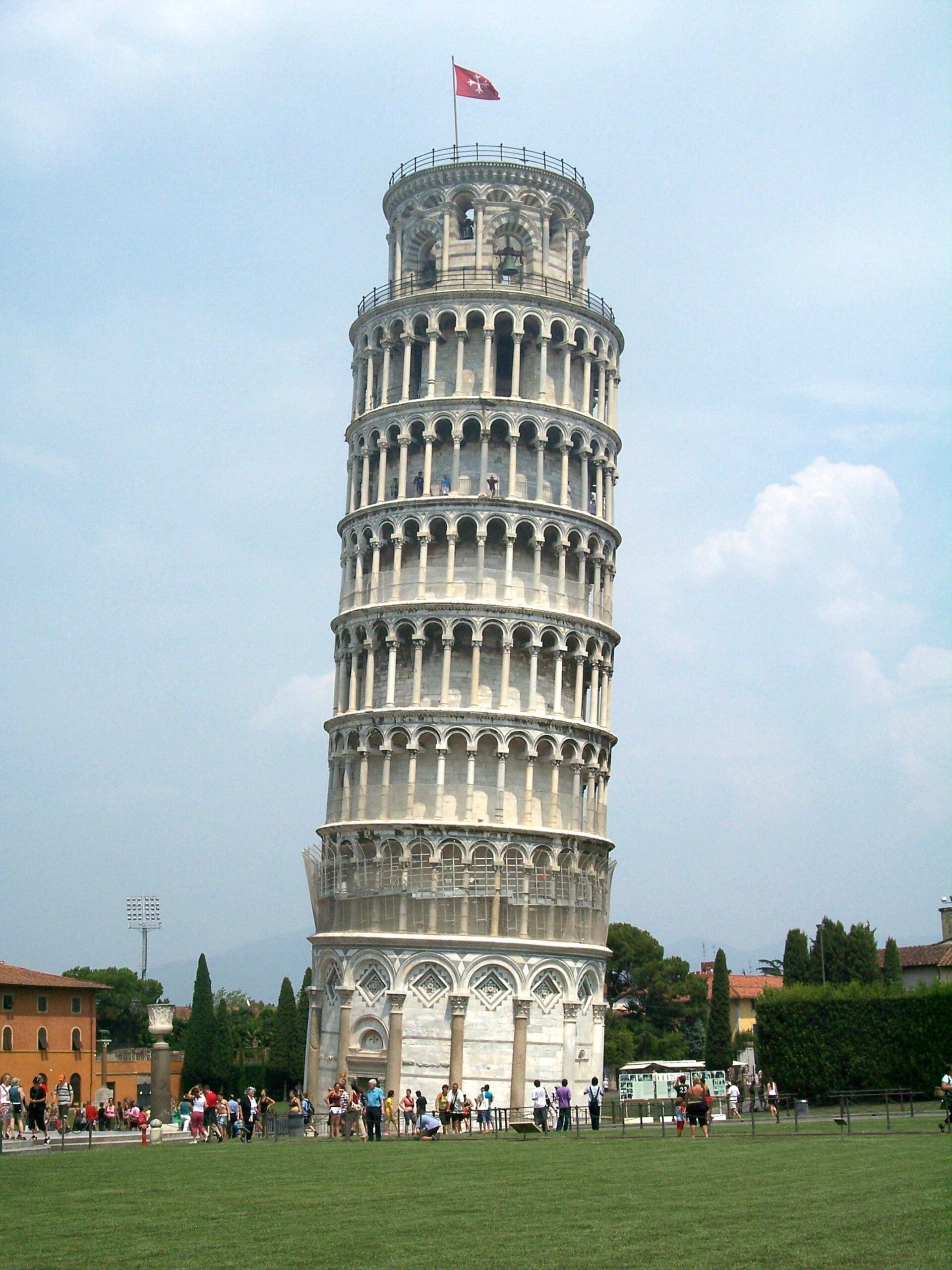 the image shows the leaning tower of pisa, a tall, cylindrical white marble bell tower with a noticeable tilt. it stands against a cloudy sky, surrounded by a lawn and tourists. a red flag flies atop the tower. the image shows the leaning tower of pisa, a tall, cylindrical white marble bell tower with a noticeable tilt. it stands against a cloudy sky, surrounded by a lawn and tourists. a red flag flies atop the tower.