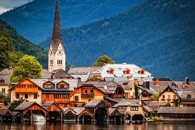a picturesque lakeside village with charming wooden houses and a prominent church with a tall steeple, set against a backdrop of lush green hills and misty mountains.