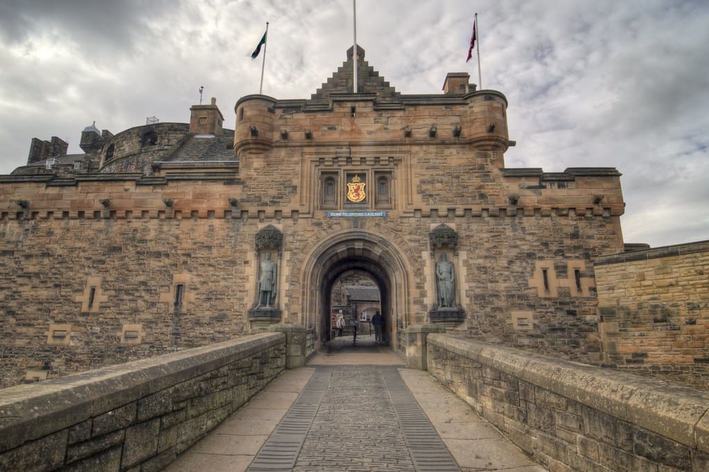 a wide stone pathway leads to the entrance of an ancient castle with an arched gate, flanked by stone walls. the castle features a heraldic emblem above the entrance and flags flying on top. the sky is overcast.